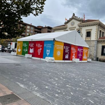 Carpa con gráficos coloridos de los ODS en un espacio público, junto a un edificio.