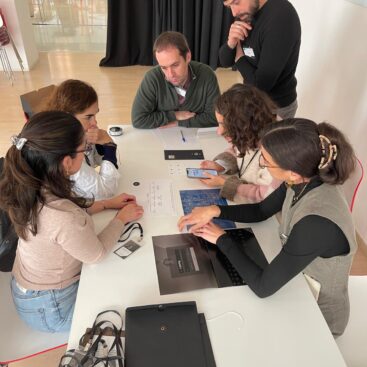 Grupo de personas sentadas alrededor de una mesa, interactuando con dispositivos móviles y documentos.
