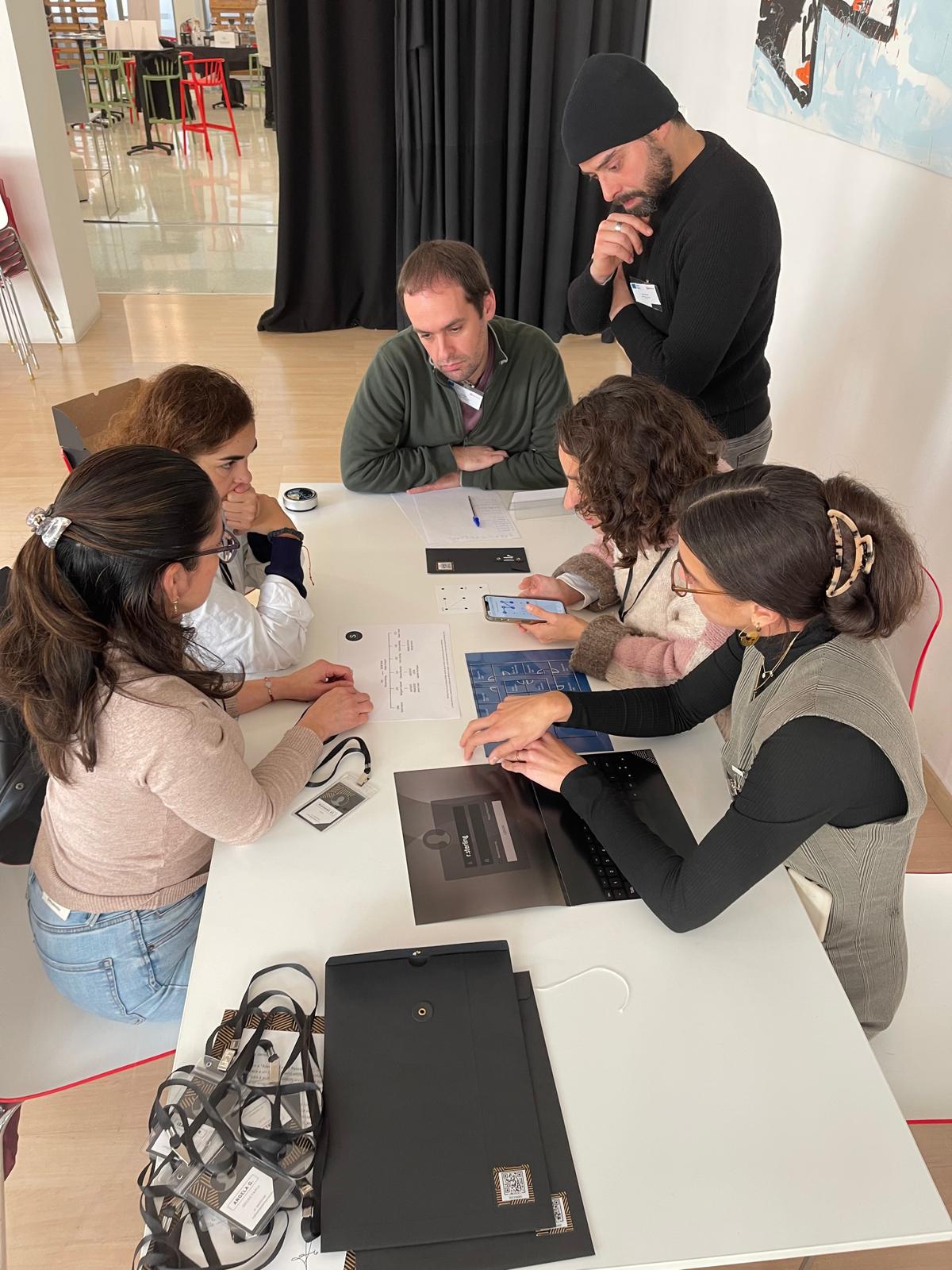 Grupo de personas sentadas alrededor de una mesa, interactuando con dispositivos móviles y documentos.
