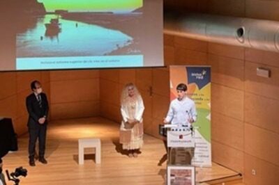 Tres personas en un escenario, una presenta mientras las otras dos escuchan. Fondo con proyección.