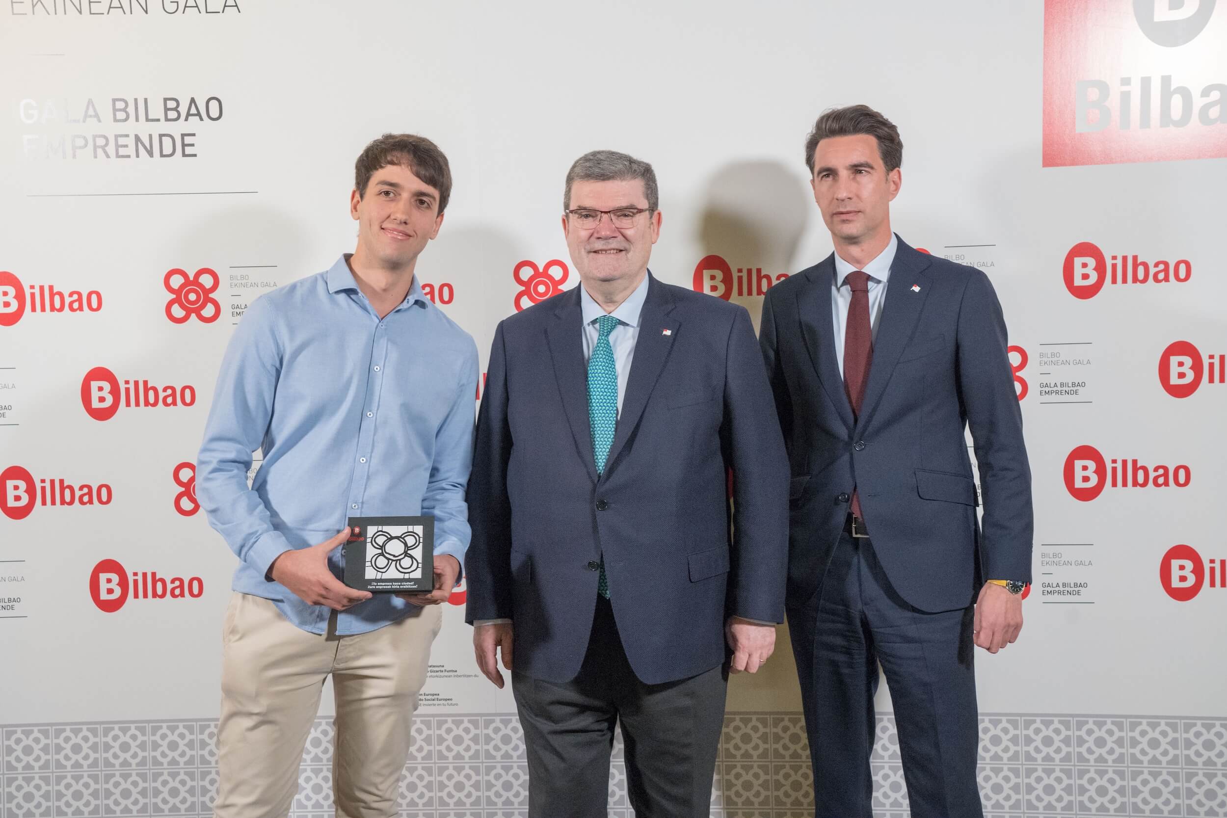 Tres personas posan en un evento, sosteniendo un premio, con fondo de logos de Bilbao.