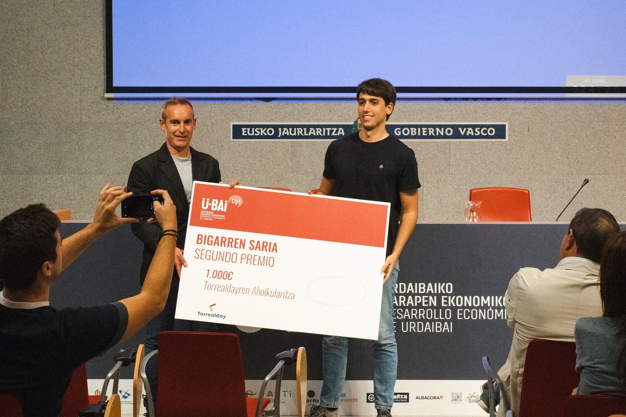 Hombre sostiene un gran cheque en un evento, mientras otro hombre lo observa.