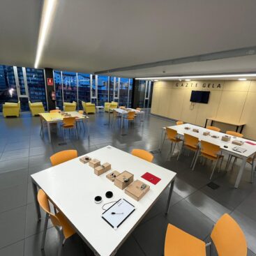 Espacio interior con mesas y sillas amarillas, ventanas grandes y vista urbana.