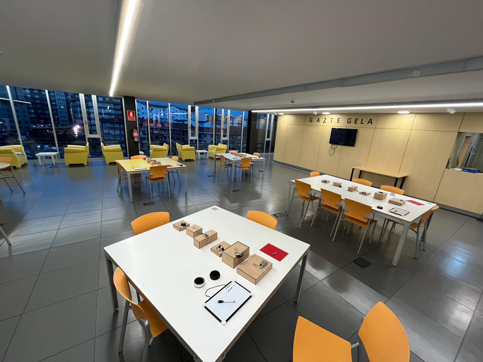Espacio interior con mesas y sillas amarillas, ventanas grandes y vista urbana.